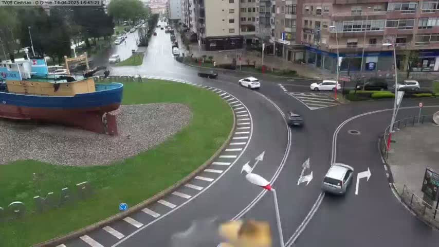 A large blue and yellow boat sits in a grassy roundabout with several cars navigating the wet roadway around it.
