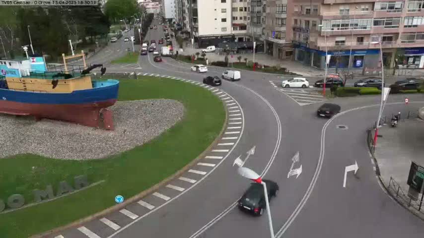 A blue and yellow fishing boat sits on a grassy roundabout next to a multi-lane roadway with several cars navigating the intersection under overcast skies.