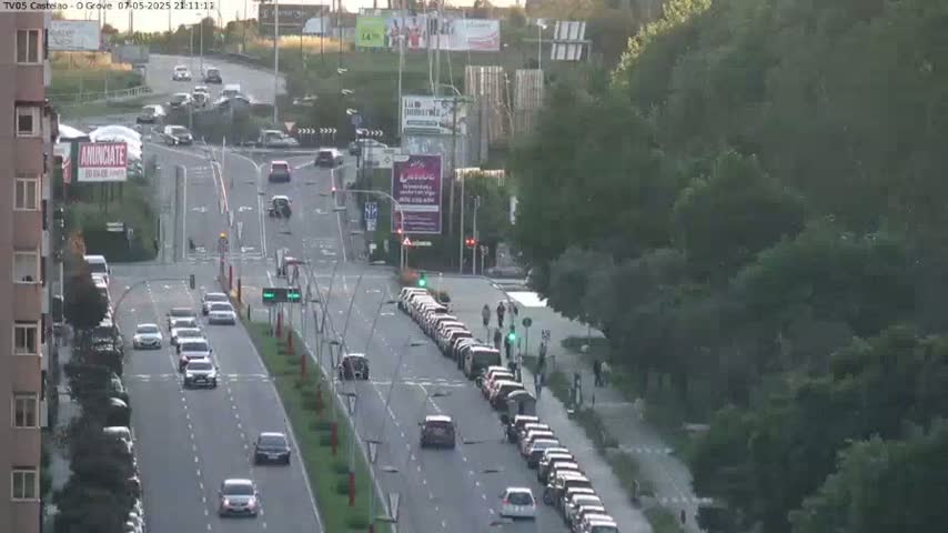 A busy multi-lane road with cars driving in both directions, under a partly sunny sky, is surrounded by lush green trees and buildings.