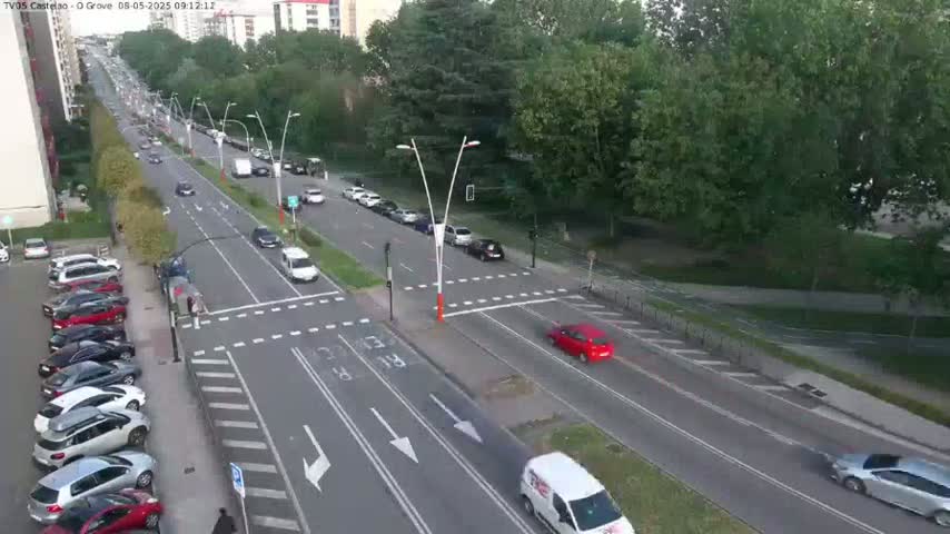 A sunny day shows a multi-lane road with cars driving in both directions, parked cars lining the side of the road, and a green park next to the road.