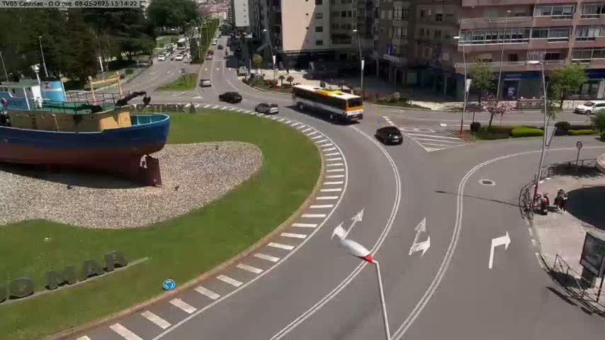 A large model boat sits in a grassy roundabout, surrounded by a sunny day and several cars and a bus navigating the curved roadway.