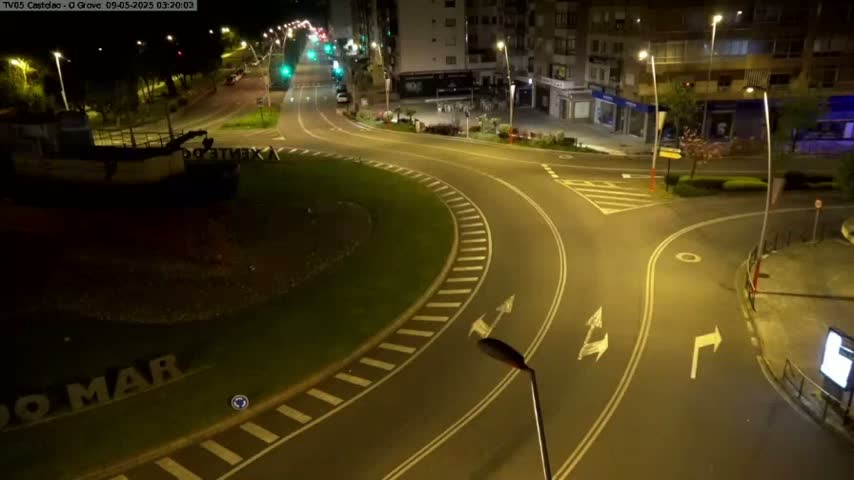 An empty roundabout at night, illuminated by streetlights, is surrounded by buildings and some greenery.