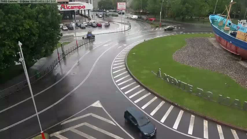 A wet roundabout with a few cars, surrounded by green space and a large ship sculpture, is seen on a rainy day.