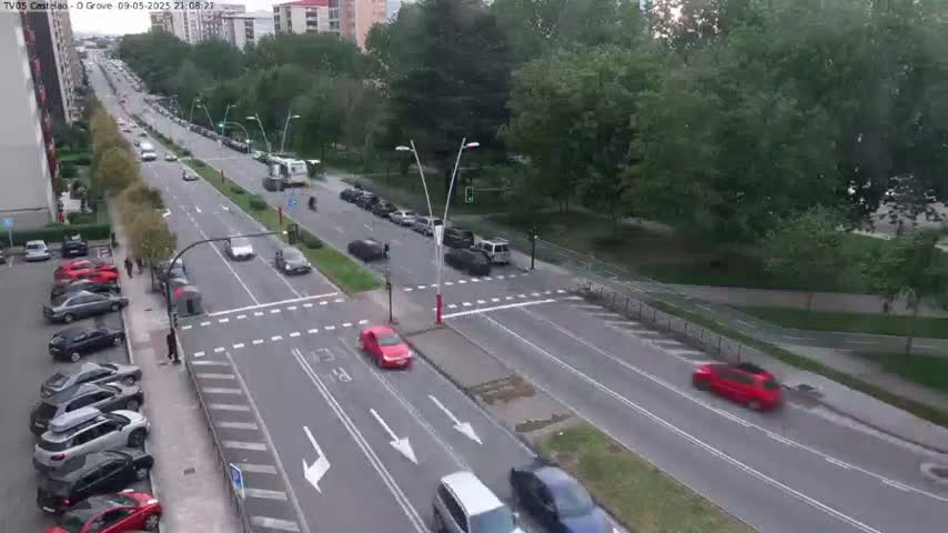 A high-angle view of a city street with several cars driving on a multi-lane road that intersects with another road, surrounded by lush green trees under a partly cloudy sky.