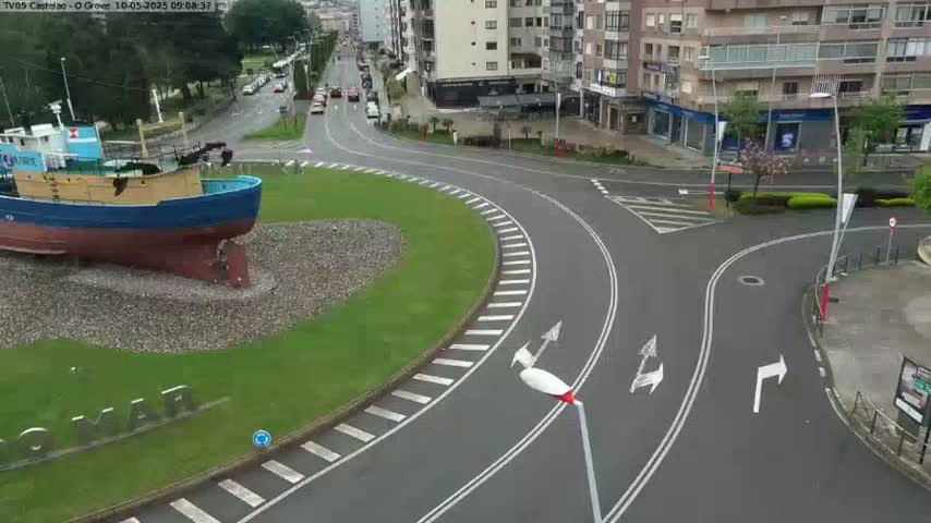 A blue and yellow fishing boat sits in a grassy roundabout at a road intersection, with several apartment buildings visible in the background under a partly cloudy sky.