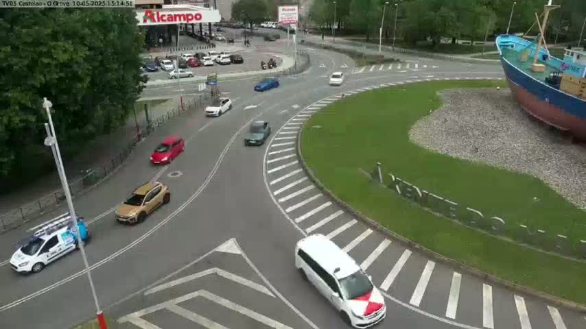 Several cars are driving around a roundabout next to a large ship, on a partly sunny day.