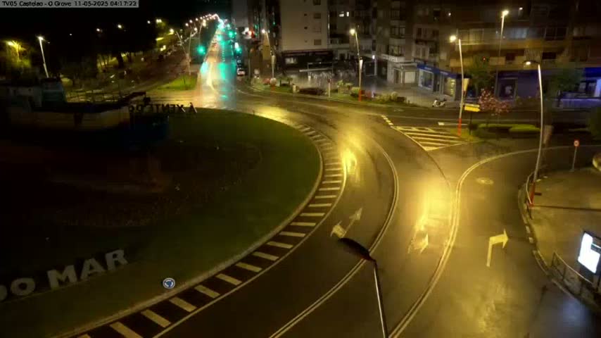 A wet roundabout at night is illuminated by streetlights, with vehicles visible on the roads leading to it.