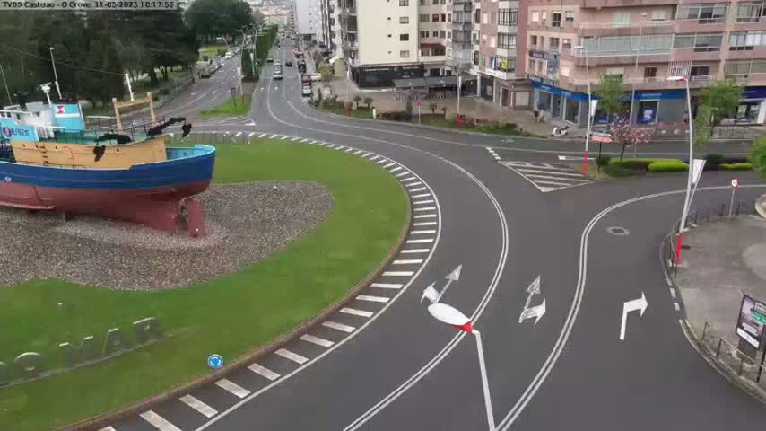 A blue and brown fishing boat sits on the grass next to a roundabout in a town, with several cars visible on the roads on a cloudy day.