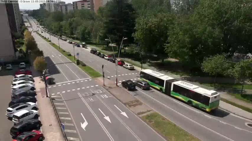 A sunny day shows a multi-lane road with cars parked along the side and several cars and a bus traveling on the road, which is surrounded by green trees.