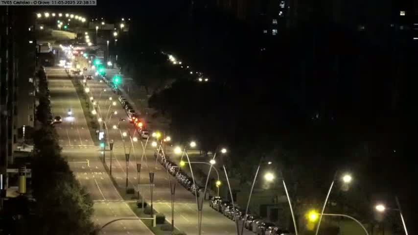 A long, well-lit road at night with cars parked along one side and cars driving on the other, under a clear, dark sky.