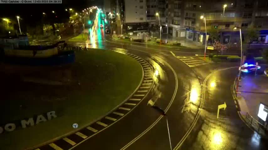 A wet, nighttime roundabout scene shows a portion of a town, with vehicles driving on the road and a boat visible near the roundabout.