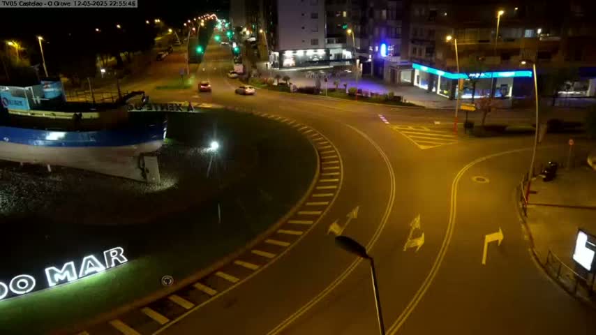A nighttime aerial view shows a roundabout with a few cars, a large boat sculpture, and buildings in the background under artificial light.