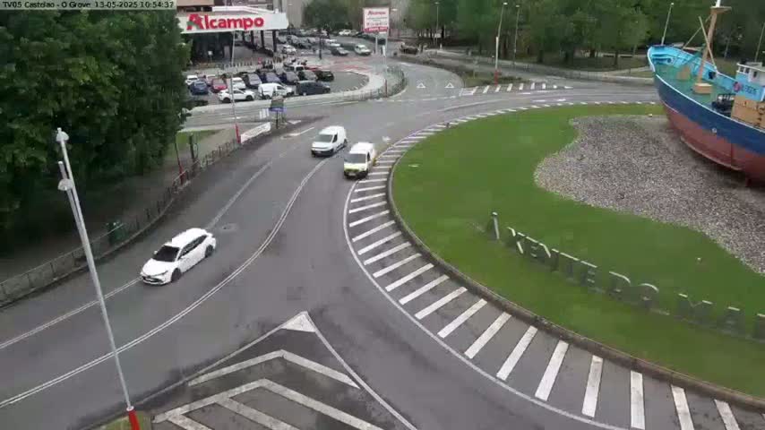 A roundabout with several cars and vans, next to a large ship, on a partly cloudy day.