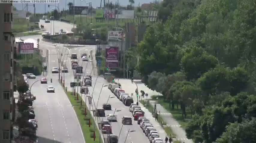 A sunny day reveals a multi-lane road with moderate traffic, flanked by lush green trees and buildings.