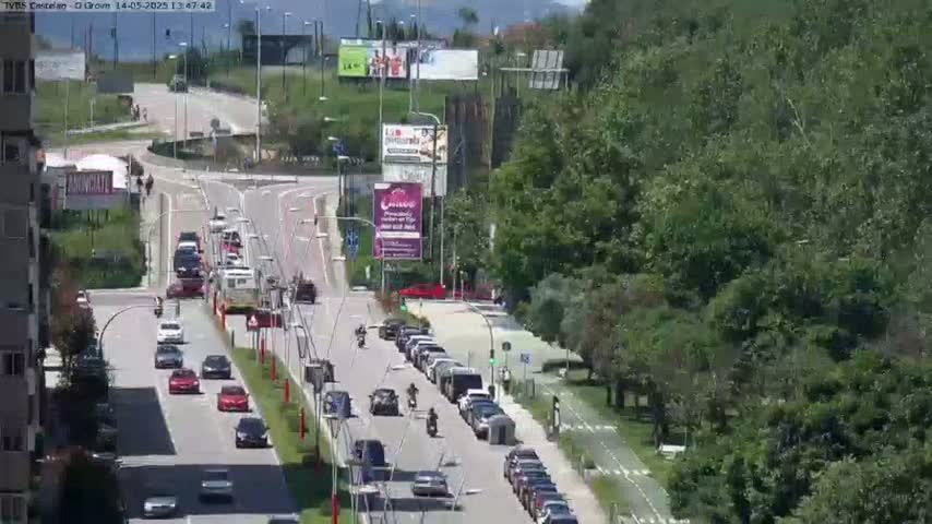 A sunny day reveals a multi-lane road with moderate traffic, flanked by lush green trees and billboards, leading to a highway overpass.