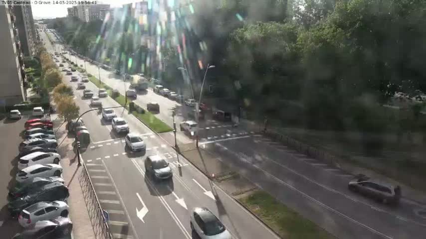 A sunny day shows a multi-lane road with cars driving in both directions, parked cars lining a street, and lush green trees along the roadside.