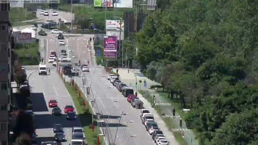 A sunny day shows a multi-lane road with cars driving in both directions, bordered by green trees and buildings.