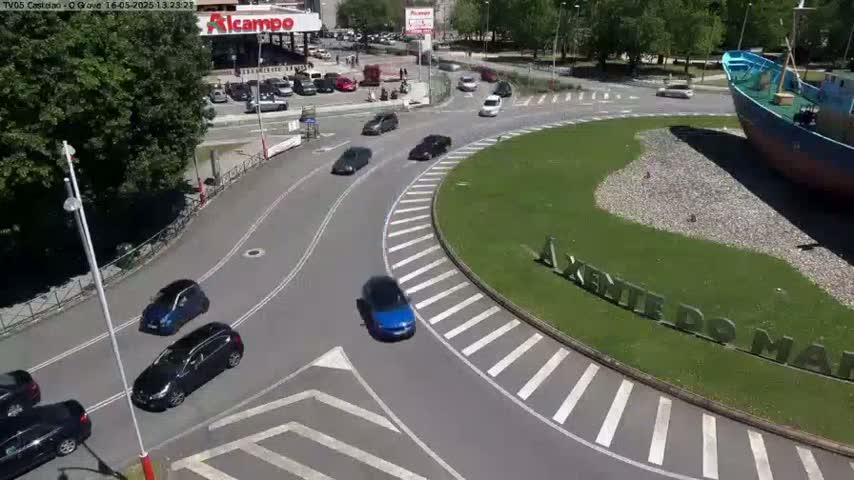 Several cars are driving around a roundabout on a sunny day, near a large boat sculpture in a grassy area.