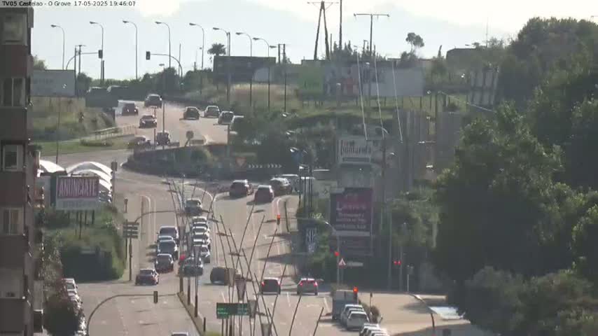 A sunny day shows a multi-lane road with moderate traffic, curving around a roundabout surrounded by green vegetation and buildings.