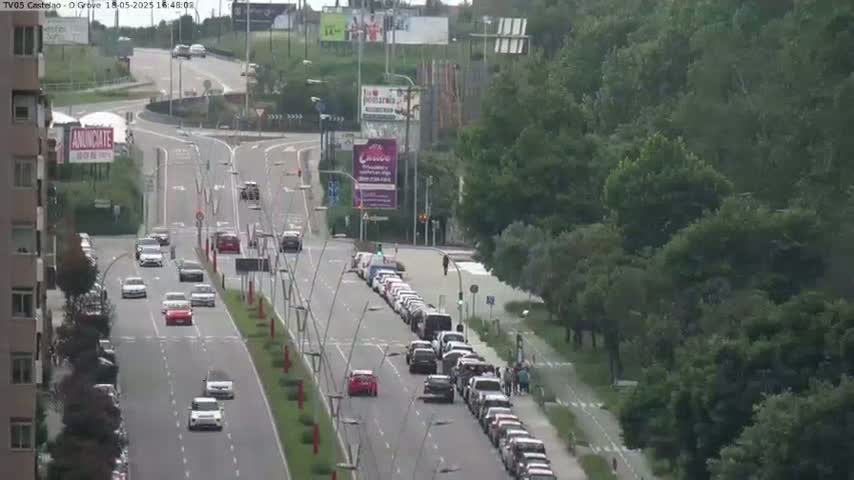 A multi-lane road with moderate traffic flows through a green, tree-lined area under a partly sunny sky.