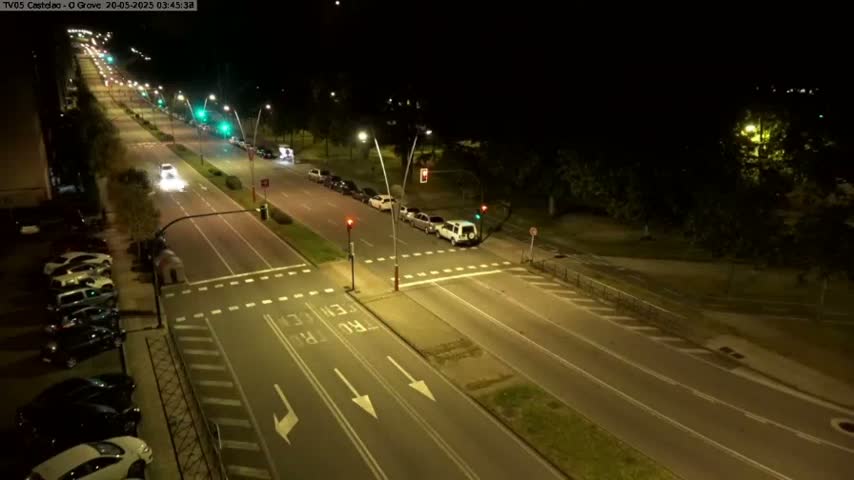 A nighttime, high-angle view of a multi-lane road intersection with cars parked along the sides and a few cars driving on the road under artificial light.