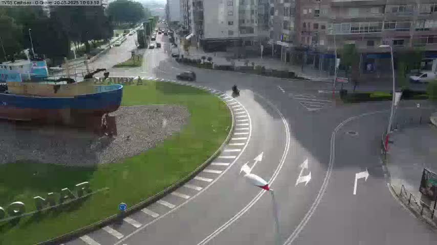 A model of a fishing boat sits in a grassy roundabout next to a roadway with several cars and a motorcycle, under sunny conditions.