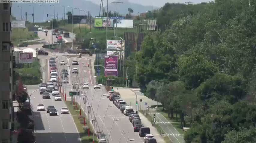 A sunny day shows a multi-lane road with moderate traffic flowing through a green, tree-lined area.