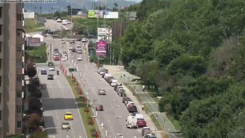 A sunny day reveals a multi-lane road with moderate traffic, flanked by lush green trees and a tall building on one side.