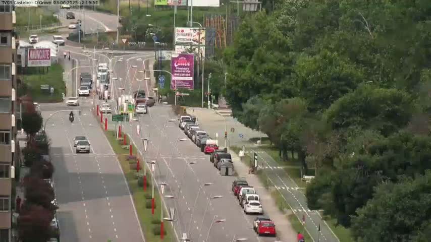 A sunny day reveals a multi-lane road with light traffic, lined with lush green trees and bordered by buildings.