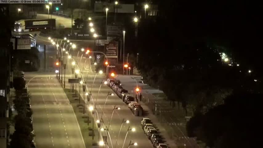 A nighttime, well-lit roadway shows several cars parked along the sides of the road, with more cars driving down the center, and a large truck visible in the distance.