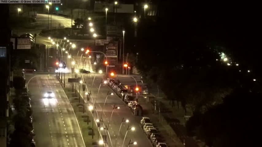 A wet, brightly lit highway at night shows several cars driving in both directions, with parked cars lining the side of the road.
