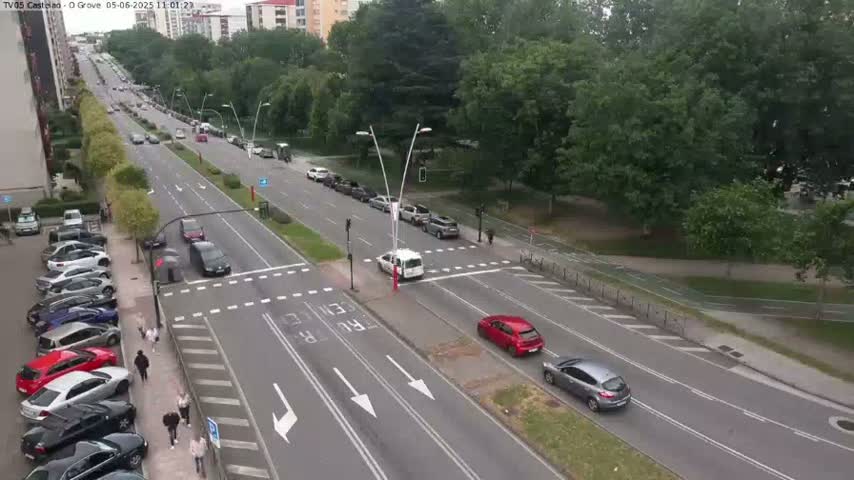 An aerial view shows a city street with several cars driving in both directions, pedestrians walking on the sidewalk, and parked cars along the side of the road, under an overcast sky.