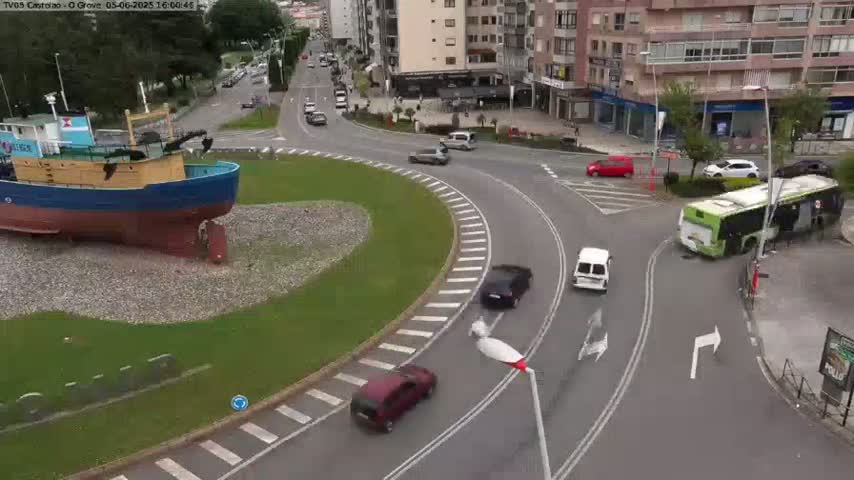 A large, blue and yellow boat sits in a grassy roundabout surrounded by a multi-lane road with several cars and a bus navigating the curves on an overcast day.