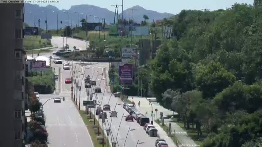A sunny day reveals a multi-lane road with moderate traffic passing through a landscape with lush green trees and buildings, some billboards visible in the distance.