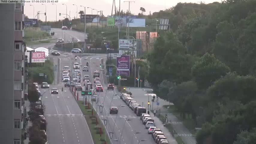 A multi-lane road with moderate traffic flows through a cityscape under a partly cloudy, twilight sky, with a large swath of green trees visible to the right.
