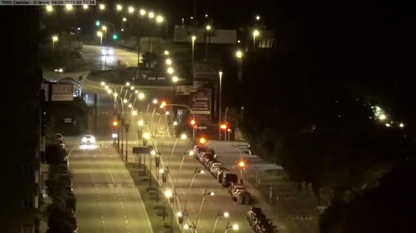 A nighttime, well-lit roadway with several cars driving, shows a roadway intersection with some parked cars on the right side.