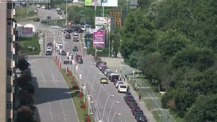 A sunny day shows a multi-lane road with cars driving in both directions,  lined with parked cars along the sides, and bordered by lush green trees.