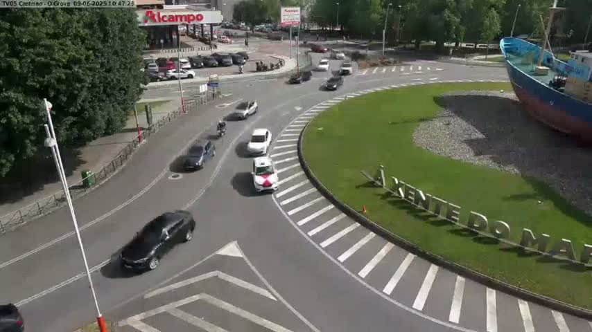 A sunny day shows several cars navigating a roundabout around a large boat on land, next to a grassy area with lettering.
