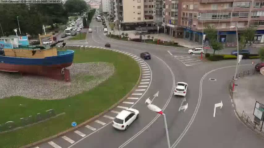 A large model of a fishing boat sits in a grassy roundabout next to a multi-lane road intersection with several cars visible, under partly cloudy skies.