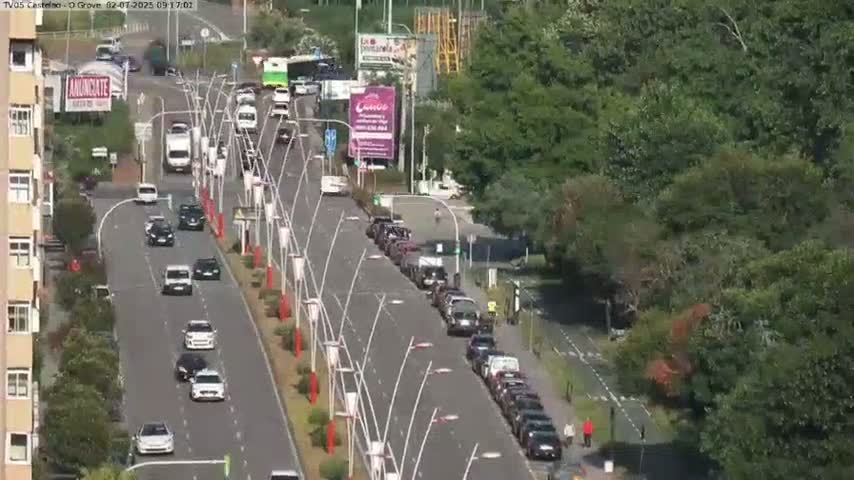 A roadway with numerous cars driving in both directions is bordered by lush green trees and buildings under a sunny sky.
