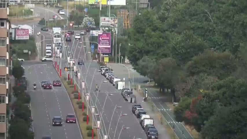 A multi-lane road with cars and a motorbike, bordered by green trees and a bicycle path, under a partly cloudy sky.