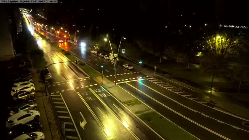 An elevated view captures a multi-lane urban road and intersection at night, with wet surfaces brightly reflecting streetlights and vehicle lights under rainy conditions.