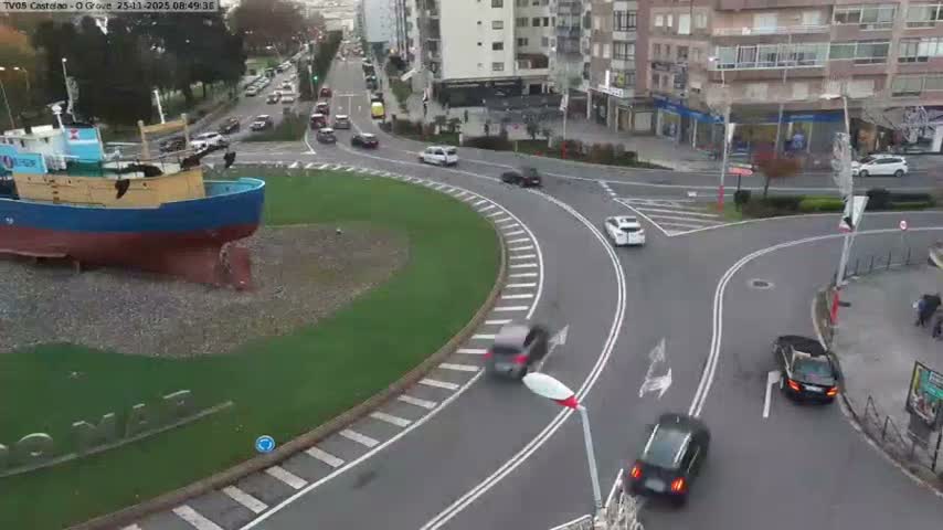 On a cloudy day, a large blue and red boat sits prominently in the grassy center of a busy urban roundabout, surrounded by vehicles and city buildings.