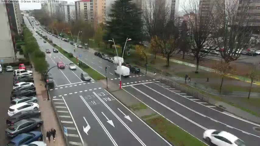 An elevated view captures a wide city street on a damp, overcast day, featuring active vehicle traffic, parked cars, pedestrians on sidewalks, green median strips, and apartment buildings in the background.