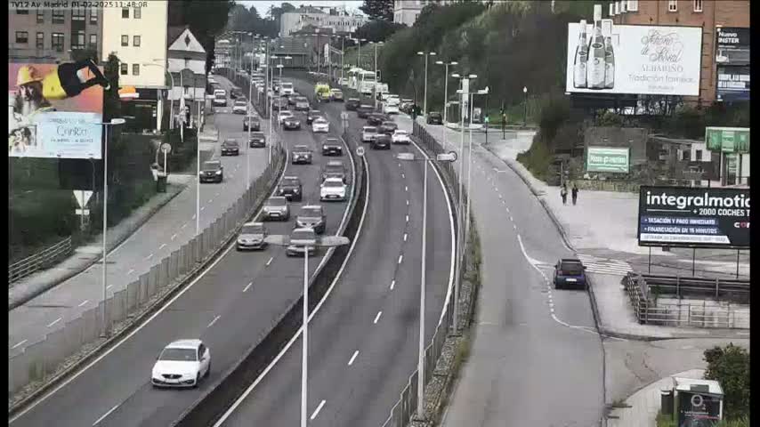 A multi-lane highway with moderate traffic flows under overcast skies, alongside a pedestrian walkway and various buildings.