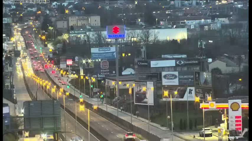 A multi-lane highway with moderate evening traffic flows past numerous billboards and buildings, under a dark, possibly overcast sky.