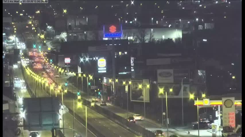 A nighttime aerial view shows a busy highway with heavy traffic under wet conditions, surrounded by illuminated buildings and streetlights.