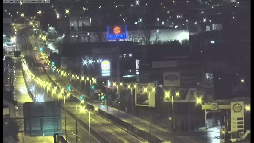 A nighttime view of a rain-slicked highway with illuminated streetlights and buildings, including a Suzuki sign.