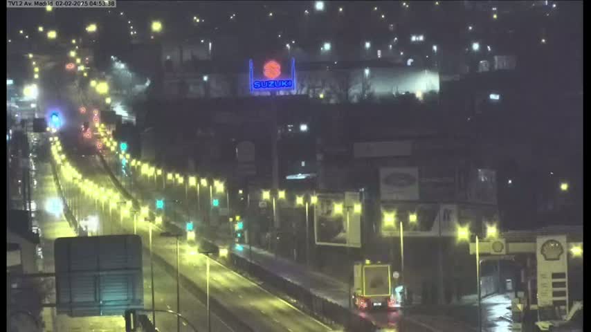 A rain-slicked highway at night is illuminated by streetlights and the headlights of cars, with a large Suzuki sign visible in the background.