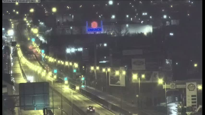 A rain-slicked highway at night is lit by streetlights and the headlights of numerous vehicles, with buildings and a large illuminated Suzuki sign visible in the background.